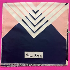 May include: A square scarf featuring a geometric pattern of navy blue, soft pink, and cream white stripes forming chevron shapes. The scarf includes a white rectangular label with the black text Nina Ricci Paris and a fringed edge at the bottom.