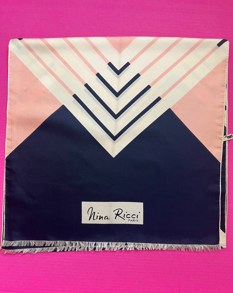 May include: A square scarf featuring a geometric pattern of navy blue, soft pink, and cream white stripes forming chevron shapes. The scarf includes a white rectangular label with the black text Nina Ricci Paris and a fringed edge at the bottom.