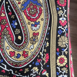 Puede incluir: Una tela textil con un patr&oacute;n de cachemira a gran escala en negro, rojo, dorado y azul. El dise&ntilde;o incluye motivos florales complejos y formas geom&eacute;tricas, con un borde vertical que muestra un patr&oacute;n repetitivo de peque&ntilde;as flores y enredaderas.