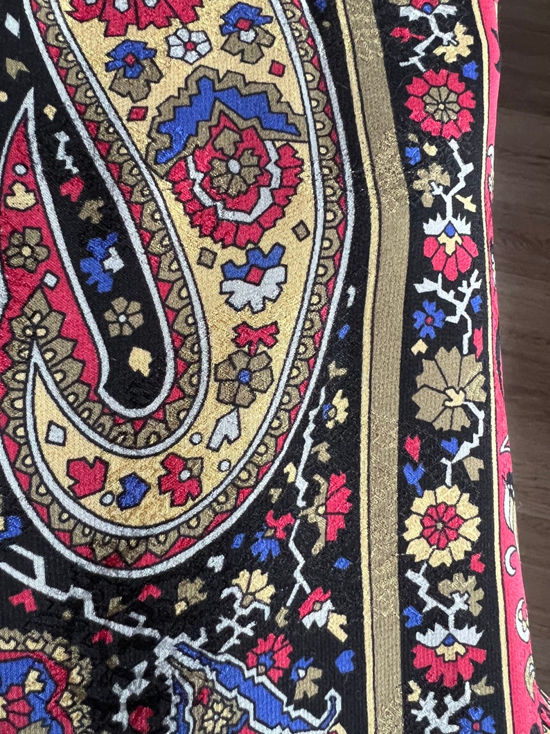 Puede incluir: Una tela textil con un patr&oacute;n de cachemira a gran escala en negro, rojo, dorado y azul. El dise&ntilde;o incluye motivos florales complejos y formas geom&eacute;tricas, con un borde vertical que muestra un patr&oacute;n repetitivo de peque&ntilde;as flores y enredaderas.