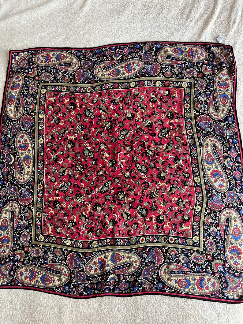 Puede incluir: Un pa&ntilde;uelo cuadrado con un panel central rojo con un denso patr&oacute;n floral negro y dorado, rodeado por un borde negro con grandes motivos de cachemira coloridos en tonos azul, amarillo y rojo. El texto Laura Biagiotti es visible.