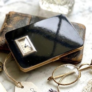 May include: A black rectangular compact with a gold-tone metal edge and an integrated analog watch face on the front. A white paper tag attached by twine reads: CIRCA 1920s ART DECO WATCH COMPACT. The compact is surrounded by a book, glasses, and perfume.