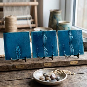 May include: Three square, deep teal ceramic tiles displayed on small metal stands. Each tile features a raised, embossed botanical pattern. Small brass plaques labelled TILE 1, TILE 2, and TILE 3 are attached to the wooden base holding the tiles.