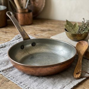 May include: A round copper frying pan with a silver-toned interior lining and a long, riveted metal handle. The pan features a warm, metallic exterior finish and a worn, functional appearance, resting on a grey linen cloth.