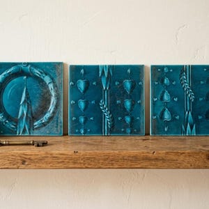 May include: Three square teal ceramic tiles featuring embossed designs. The left tile displays a circular wreath motif. The centre and right tiles feature vertical patterns of small heart shapes and leaf-like elements with a glossy finish.