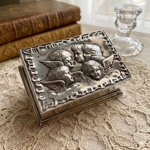 May include: A rectangular silver-toned metal box featuring a raised relief design on the lid. The lid displays four winged cherub faces arranged in a cluster, surrounded by a decorative border with a textured, scalloped edge pattern.
