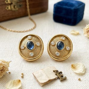 May include: A pair of round gold-tone earrings with a textured finish, each featuring a central oval blue cabochon stone surrounded by four small clear round rhinestones arranged in a cross pattern on the face of the circular metal disc.
