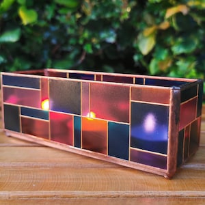 Könnte beinhalten: Ein rechteckiger Kerzenhalter mit einem Buntglasdesign. Er hat einen Kupferrahmen und ein Mosaik aus farbigen Glasquadraten in Rot-, Blau- und Lilatönen. Angezündete Kerzen im Inneren erzeugen ein warmes Leuchten. Die Länge beträgt ca. 20 cm.