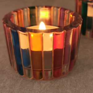 May include: Two glass candle holders with lit candles. The holders are round with vertical stripes in various colours, including blue, red, and yellow. The candles are white and provide a warm glow.