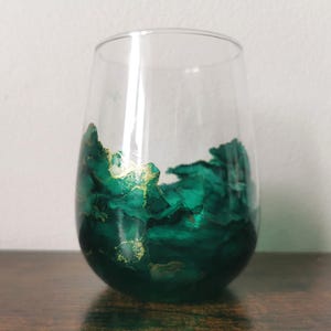 May include: A stemless wine glass with an emerald green and gold abstract design. The glass has a clear upper section and a painted lower section, creating a unique decorative effect. The design resembles flowing ink.