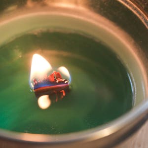 Puede incluir: Una vela verde encendida en un recipiente plateado. La llama es brillante, con una mecha roja y azul. La cera de la vela es de color verde oscuro y el recipiente es redondo.