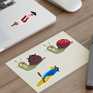 May include: Four stickers featuring cartoon snails and a parrot wearing pirate hats. The snails are green and brown with black eyes and a skull and crossbones on their shells. The parrot is blue with a yellow beak and a black eye patch. The stickers are on a light brown wooden surface.