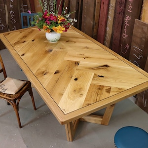 Op de afbeelding: Een houten eettafel met een visgraatmotief bovenblad en een donkerhouten rand. De tafel heeft een eenvoudig, modern ontwerp met een rechthoekig blad en een sokkelonderstel. De tafel wordt getoond in een werkplaats met een stoel en een kruk.