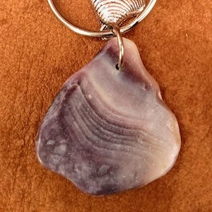 Puede incluir: Un llavero plateado con un mosquetón, un pequeño dije de concha y un colgante grande de concha morada y blanca de forma irregular. El llavero se muestra sobre un fondo marrón.