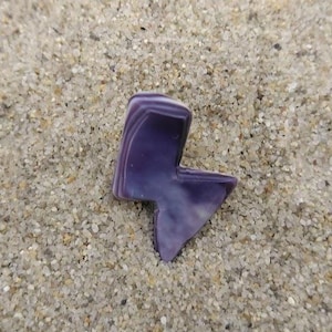 Puede incluir: Una piedra morada y blanca con forma de rayo descansa sobre arena. La piedra tiene un diseño en capas con tonos de morado y blanco. El fondo es un primer plano de arena.