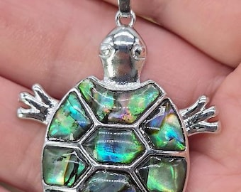 Pode incluir: Um colar com pingente de tartaruga prateada. A carapaça da tartaruga é incrustada com peças iridescentes de concha de abalone em tons de verde, azul e roxo. O pingente é preso a uma delicada corrente de prata, criando uma joia única.