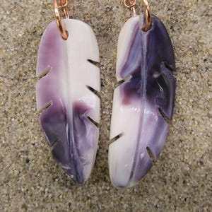 Puede incluir: Un par de pendientes con forma de pluma en tonos morado y blanco, con ganchos de color cobre. Los pendientes están sobre un fondo arenoso. Una joya única.