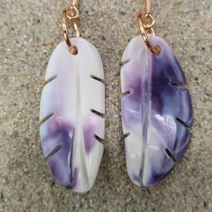Puede incluir: Un par de pendientes con diseño de pluma. Los pendientes son blancos con vetas moradas y tienen ganchos de color oro rosa. Se exhiben sobre una superficie arenosa.
