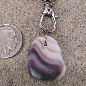 Puede incluir: Llavero con un colgante de piedra pulida de forma ovalada. La piedra presenta bandas arremolinadas de blanco, lavanda y morado oscuro. El llavero tiene un cierre y un anillo plateados. Una moneda antigua está al lado del colgante. El fondo es arena.