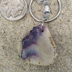 Puede incluir: Un llavero plateado con un dije de cola de sirena y un colgante de concha morada y blanca. Una moneda de plata está al lado del llavero. Los artículos están sobre una superficie arenosa.