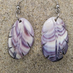 Puede incluir: Un par de pendientes artesanales con diseño de concha. Los pendientes presentan una combinación de colores blanco y morado con un patrón de hoja tallada. Cuelgan de ganchos plateados.