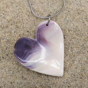 May include: A heart-shaped pendant necklace with a silver chain. The pendant is a mix of white and purple, with a glossy finish. The necklace is resting on a sandy surface, creating a beachy aesthetic.