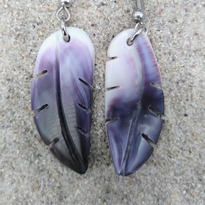 Puede incluir: Un par de pendientes con forma de pluma. Cada pendiente presenta un diseño tallado con un degradado de colores que va del blanco a los tonos púrpura y negro. Los pendientes tienen ganchos plateados y se muestran sobre una superficie arenosa. Miden unos 5 cm.