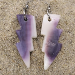 Puede incluir: Un par de pendientes con forma de rayo. Los pendientes son blancos y morados con ganchos plateados. Los pendientes se exhiben sobre una superficie arenosa.