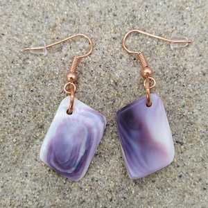 Puede incluir: Un par de pendientes con colgantes rectangulares veteados en morado y blanco. Los pendientes tienen ganchos de color cobre y se muestran sobre una superficie arenosa. Son una pieza de joyería.