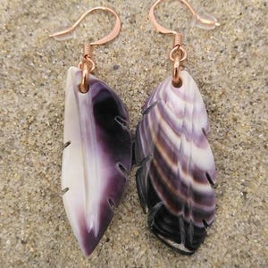 Puede incluir: Un par de pendientes en forma de pluma. Cada pendiente está hecho de una concha pulida, con un degradado de tonos blancos, morados y negros. Los pendientes tienen ganchos para las orejas de color cobre, sobre un fondo arenoso.