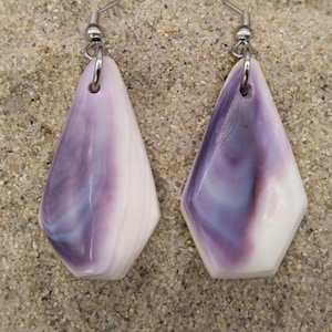 Puede incluir: Un par de pendientes colgantes con forma geométrica. Cada pendiente presenta un diseño veteado blanco y morado, con un gancho plateado. Los pendientes se muestran sobre una superficie arenosa.