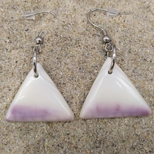 Puede incluir: Un par de pendientes triangulares blancos y lavanda con ganchos plateados. Los pendientes se exhiben sobre una superficie arenosa, sugiriendo un tema de playa o costero. Los pendientes tienen un acabado brillante.