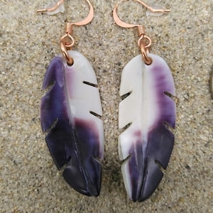 Puede incluir: Un par de pendientes con forma de pluma con un degradado de blanco a morado oscuro. Los pendientes tienen ganchos de color cobre y están sobre una superficie arenosa. El diseño de la pluma tiene detalles recortados.