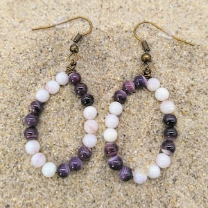 Puede incluir: Un par de pendientes de cuentas de concha púrpura y blanca con alambre y ganchos de bronce. Las cuentas son redondas y tienen un degradado de color de púrpura a blanco.