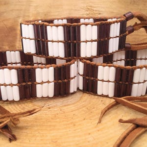 Puede incluir: Tres pulseras de cuentas marrones y blancas con borlas de cuero. Las pulseras están hechas con cuentas pequeñas y rectangulares en un patrón de cuadros.