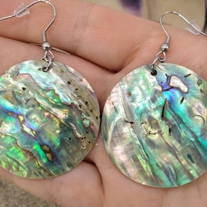 May include: A pair of iridescent abalone shell earrings. Each earring has a round, polished shell with a unique pattern of greens, blues, and purples. The earrings have silver-coloured fish hook ear wires.