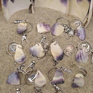 Puede incluir: Una colección de llaveros con piedras naturales pulidas en tonos morados y blancos, unidos a anillos y cierres de metal plateado. Los llaveros están colocados sobre arena, con grandes conchas marinas en el fondo.