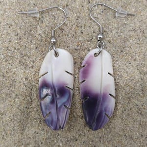 Puede incluir: Un par de pendientes en forma de pluma. Cada pendiente es blanco en la parte superior, pasando a un morado intenso en la parte inferior. Los pendientes tienen un diseño de pluma tallada con pequeños cortes angulados. Cuelgan de ganchos y postes plateados.