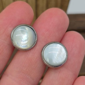 May include: A pair of round stud earrings with a silver-coloured border and iridescent, white centres. The earrings are displayed against a neutral background, highlighting their simple, elegant design. A classic jewellery item.