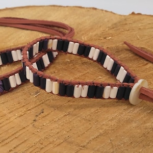 Puede incluir: Un brazalete de cuero marrón con cuentas blancas y negras. El brazalete tiene un largo corbatín de cuero marrón.