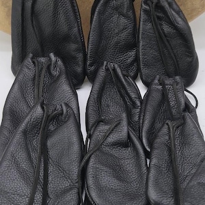 May include: A collection of seven black drawstring pouches made of textured material. Each pouch features a gathered top secured with a thin black cord. The pouches are arranged in two rows, displaying a consistent matte black finish and a soft, flexible structure with visible grain patterns on the surface of each individual bag.