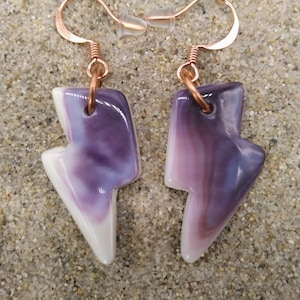 Peut inclure: Paire de boucles d'oreilles artisanales en forme d'éclair. Les boucles d'oreilles ont des crochets de couleur cuivre et une pierre marbrée violette et blanche. Les boucles d'oreilles sont présentées sur une surface sablonneuse.