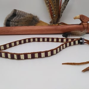 Puede incluir: Un collar de cuero marrón con cuentas blancas y moradas. El collar tiene dos borlas de cuero.