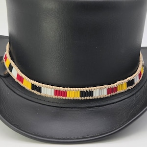 Puede incluir: Un sombrero de copa de cuero negro con una banda decorativa hecha de cordón beige tejido y pequeñas cuentas cilíndricas en un patrón repetitivo de rojo, amarillo, negro y blanco. El sombrero tiene un acabado mate y un ala ancha.