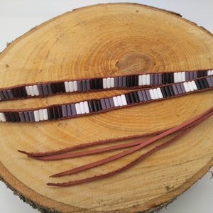 Puede incluir: Un cordón de cuero marrón con flecos de cuentas y una doble fila de cuentas de vidrio blancas, negras y moradas.