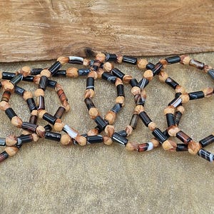 May include: Six beaded bracelets with a mix of black, brown, and tan cylindrical beads. The bracelets are arranged in a circular pattern against a textured, light brown surface and a wooden background.
