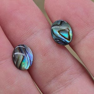 May include: Two oval-shaped abalone shell cabochons. The iridescent shells display a mix of blue, green, and silver hues. The polished stones are smooth and reflect light, showcasing their natural patterns. These cabochons are ideal for jewelry making.