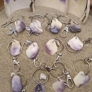 Puede incluir: Una colección de llaveros con piezas de conchas marinas pulidas en tonos morados y blancos. Cada llavero tiene un anillo y un cierre plateados. Algunos llaveros tienen dijes de estrella de mar o conchas. El fondo es arena con conchas grandes.