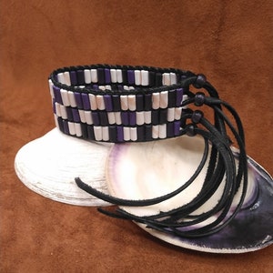 Puede incluir: Un brazalete de cuentas blancas y moradas con lazos de cuero negro. El brazalete está sobre una concha blanca.