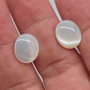 May include: Two oval, cabochon-cut gemstones with a pearly, iridescent sheen. The stones are a creamy white color and appear smooth and polished. The gemstones are likely used for jewelry making.